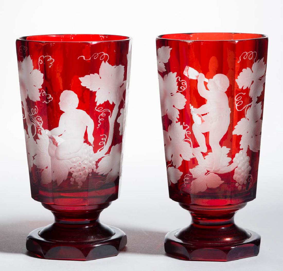 BOHEMIAN CUT AND ENGRAVED - RUBY-STAINED PAIR OF: BOHEMIAN CUT AND ENGRAVED - RUBY-STAINED PAIR OF BEAKERS / FOOTED TUMBLERS, colorless, each with designs depicting Bacchus drinking wine while standing or sitting on a grape vine, factory-polished rim
