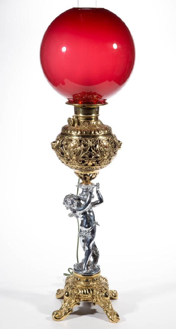 CAST-METAL FIGURAL STEM BANQUET LAMP: CAST-METAL FIGURAL STEM BANQUET LAMP, cased dark rose/ruby ball-form shade with satin interior, brass heat ring; globular-form font vase with cherub and scroll ornamentation in a brass finish, stem de