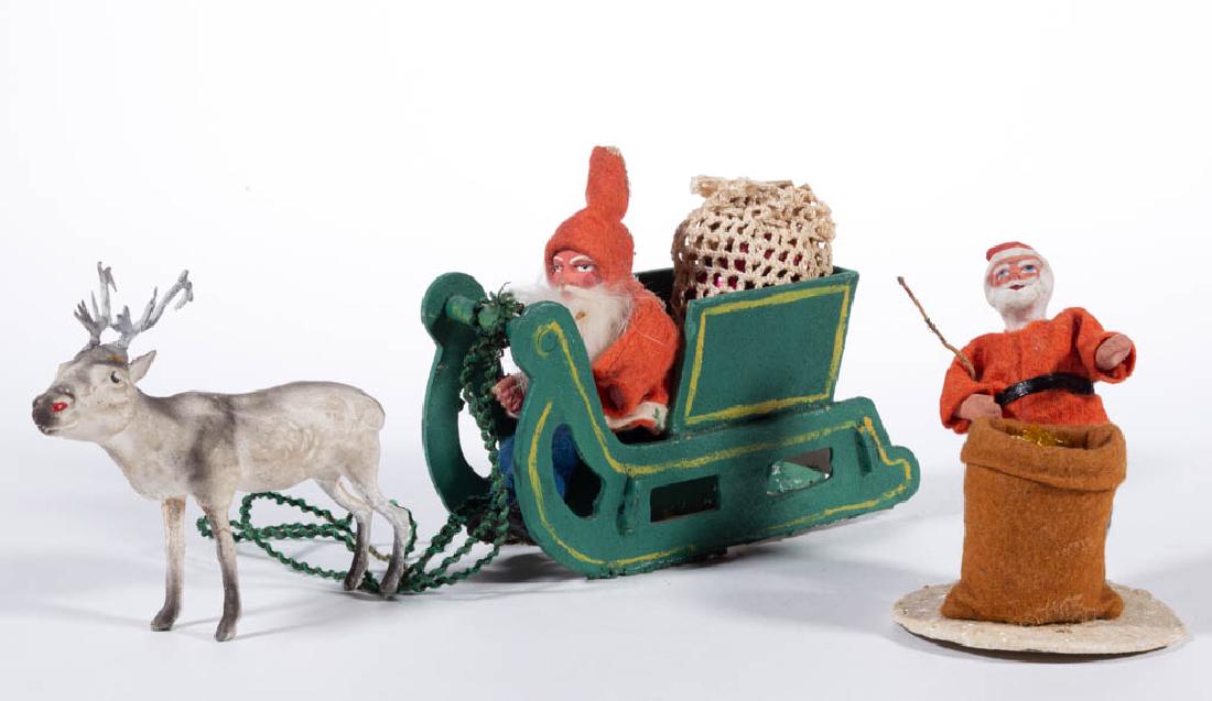 GERMAN SANTA CLAUS FIGURAL CANDY CONTAINERS, LOT OF TWO: GERMAN SANTA CLAUS FIGURAL CANDY CONTAINERS, LOT OF TWO, comprising an example standing next to a brown bag and an example sitting in a sleigh with bag. Together with a composition deer figure. Each f