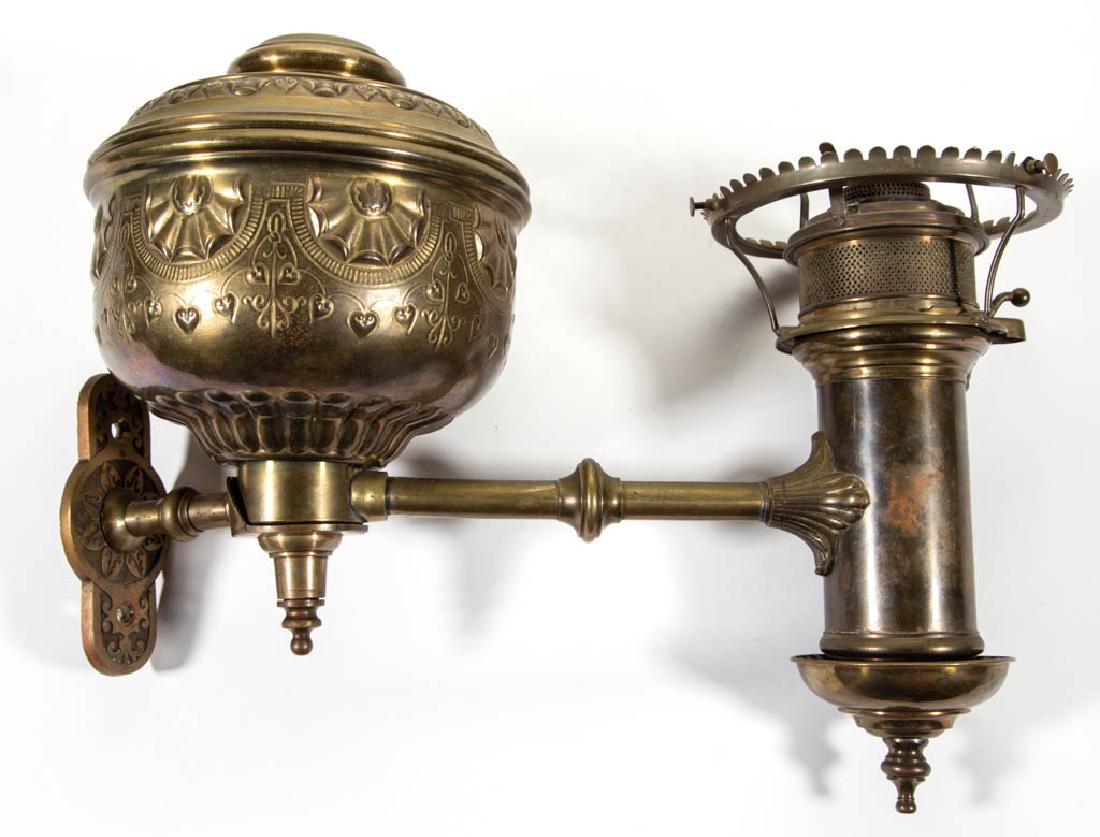 E. MILLER STAMPED BRASS WALL-MOUNTED KEROSENE STUDENT: E. MILLER STAMPED BRASS WALL-MOUNTED KEROSENE STUDENT LAMP, stamped-brass font vase with shell and hanging hearts motif, original font tank, cylindrical reservoir with period burner with correct flame