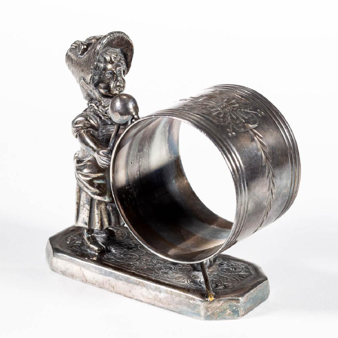 SIMPSON, HALL, MILLER & CO. KATE GREENWAY-TYPE FIGURAL: SIMPSON, HALL, MILLER & CO. KATE GREENWAY-TYPE FIGURAL SILVER-PLATED NAPKIN RING, depicting a Kate Greenway-type girl holding a napkin ring which is fastened to her by a rope, representing a drum, she