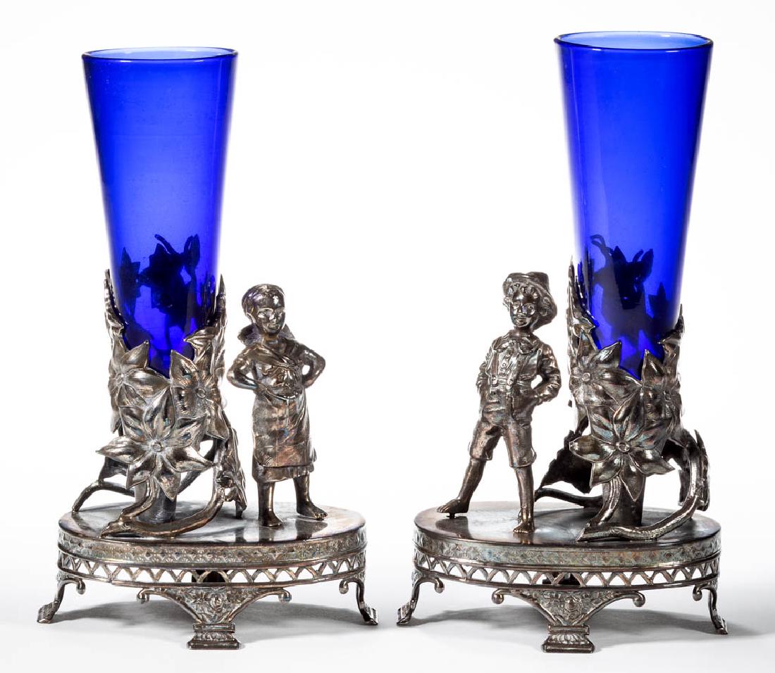 HARTFORD SILVER PLATE CO. FIGURAL SILVER-PLATED VASE: HARTFORD SILVER PLATE CO. FIGURAL SILVER-PLATED VASE PAIR, each depicting a child figure standing next to a floral vase holder with blue glass insert, oval base with flower basket-type feet. Each with