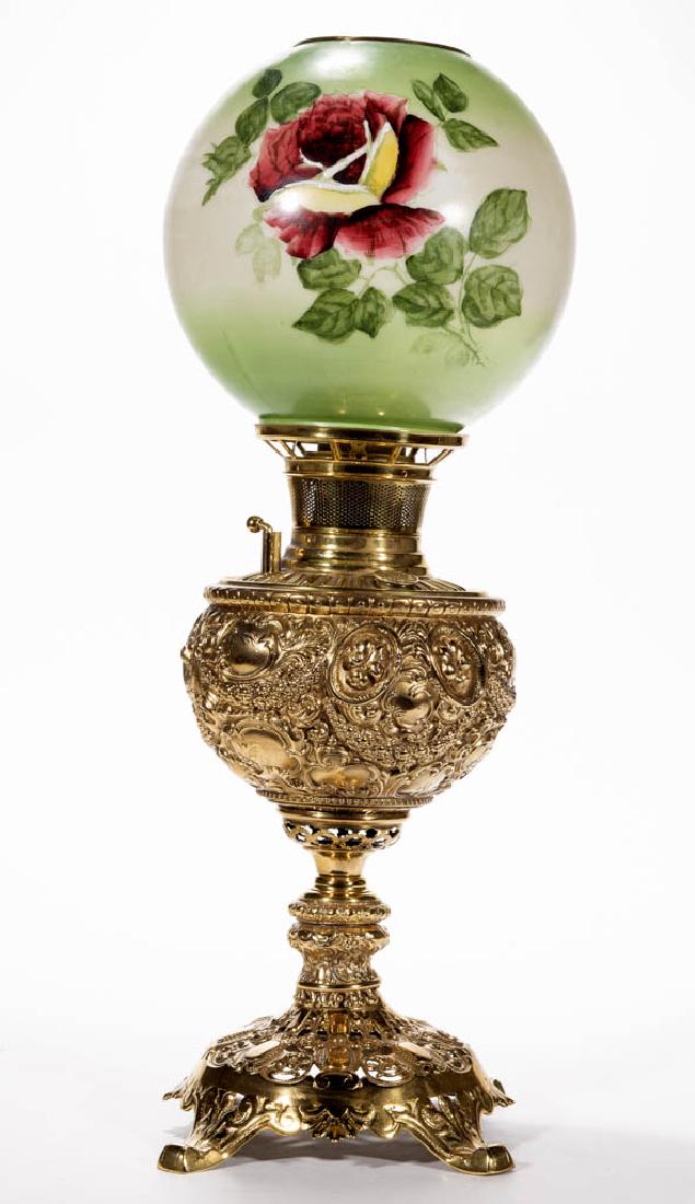 BRASS AND CAST METAL KEROSENE VASE TABLE LAMP: BRASS AND CAST METAL KEROSENE VASE TABLE LAMP, opaque white ball-form shade with green stain and polychrome rose decoration, brass heat ring, globular-form font holder with heavy ornamentation of flor