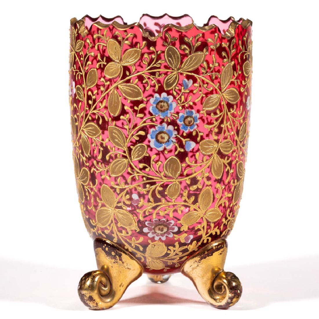 MOSER ATTRIBUTED ENAMEL-DECORATED FOOTED VASE: MOSER ATTRIBUTED ENAMEL-DECORATED FOOTED VASE, cranberry with polychrome-enamel and gilt decoration, Moorish rim with swirling floral decoration, three applied coiled feet. Bohemia, now Czech Republic