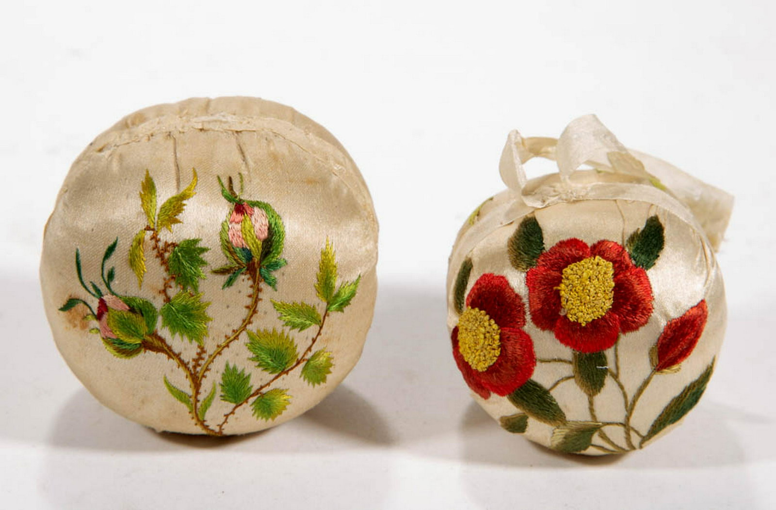 EMBROIDERED PIN-BALL CUSHIONS, LOT OF TWO: EMBROIDERED PIN-BALL CUSHIONS, LOT OF TWO, hand-stitched, each in ball form with fine embroidered floral designs on each side in polychrome thread on a white satin material ground. First half 19th cen