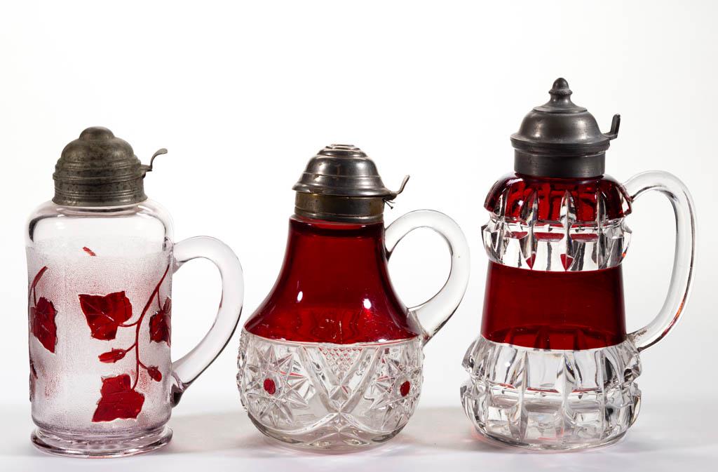 ASSORTED EAPG - RUBY-STAINED SYRUP PITCHERS, LOT OF: ASSORTED EAPG - RUBY-STAINED SYRUP PITCHERS, LOT OF THREE, colorless, comprising an Ivy in Snow, a Truncated Cube, and a Robinson' Puritan example, each with a period lid. Various makers. Fourth quart