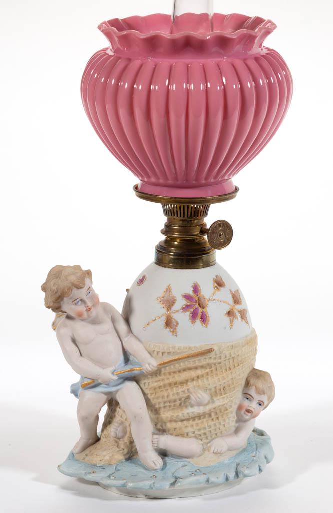 GERMAN BISQUE DOUBLE CHERUB FIGURAL MINIATURE LAMP: GERMAN BISQUE DOUBLE CHERUB FIGURAL MINIATURE LAMP, multi-colors in pastels and with gilt decoration, depicting an ovoid-form font on an overlapping leaf base, font wrapped in a burlap bag with a cher