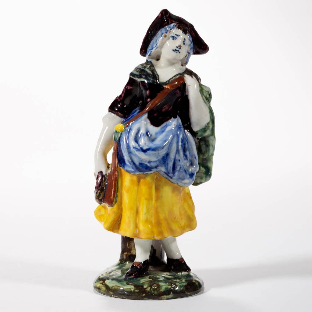 DUTCH DELFT TIN-GLAZED EARTHENWARE FIGURE: DUTCH DELFT TIN-GLAZED EARTHENWARE FIGURE, standing on a circular base, wearing a polychrome dress, tri-cornered hat with mop cap, and bag slung across shoulder. Red hand-brushed initials to base. Mid