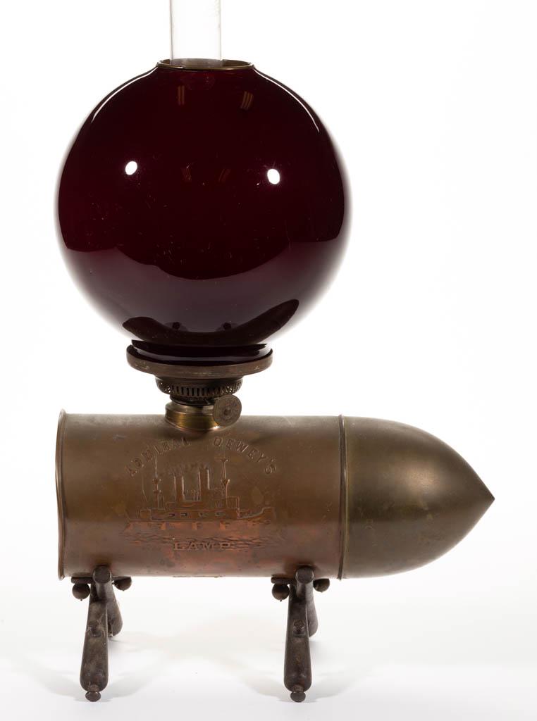 ADMIRAL DEWEY'S FIGURAL LAMP BRASS MINIATURE LAMP: ADMIRAL DEWEY'S FIGURAL LAMP BRASS MINIATURE LAMP, bullet-form font with black cast metal figural cannon feet, ship embossed on the front and "Remember The Maine" on the back, cased dark burgundy ball
