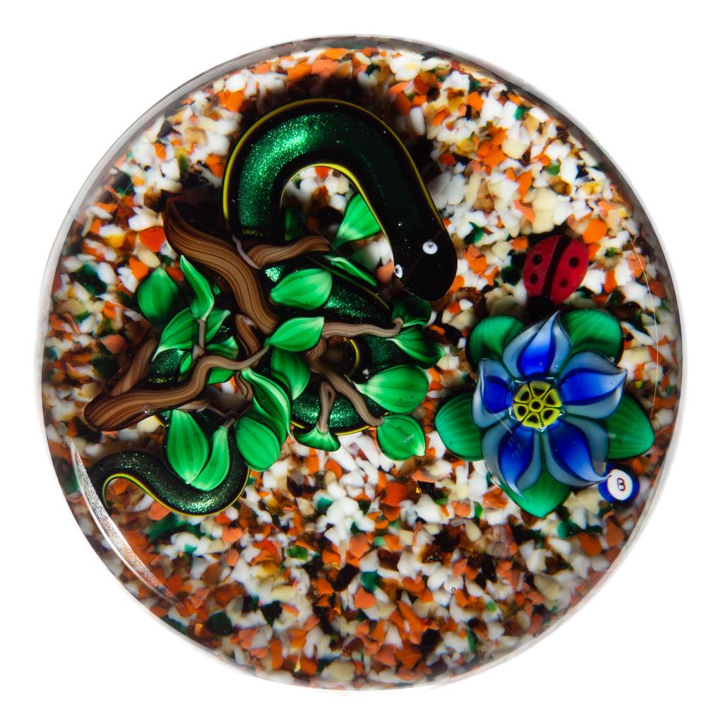BOB BANFORD SNAKE LAMPWORK STUDIO ART GLASS PAPERWEIGHT: BOB BANFORD SNAKE LAMPWORK STUDIO ART GLASS PAPERWEIGHT, colorless with a polychrome frit ground, green and yellow snake coiled under two branches with many leaves, blooming blue and white five-petal 