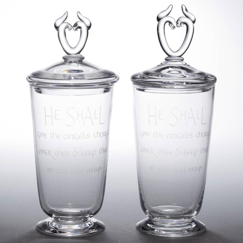STEUBEN ENGRAVED CRYSTAL PAIR OF COVERED URNS: STEUBEN ENGRAVED CRYSTAL PAIR OF COVERED URNS, colorless, each slightly tapered form with matching covers with heart-form finials, each engraved "HE SHALL/GIVE THE ANGELS CHARGE/OVER THEE & KEEP THEE/