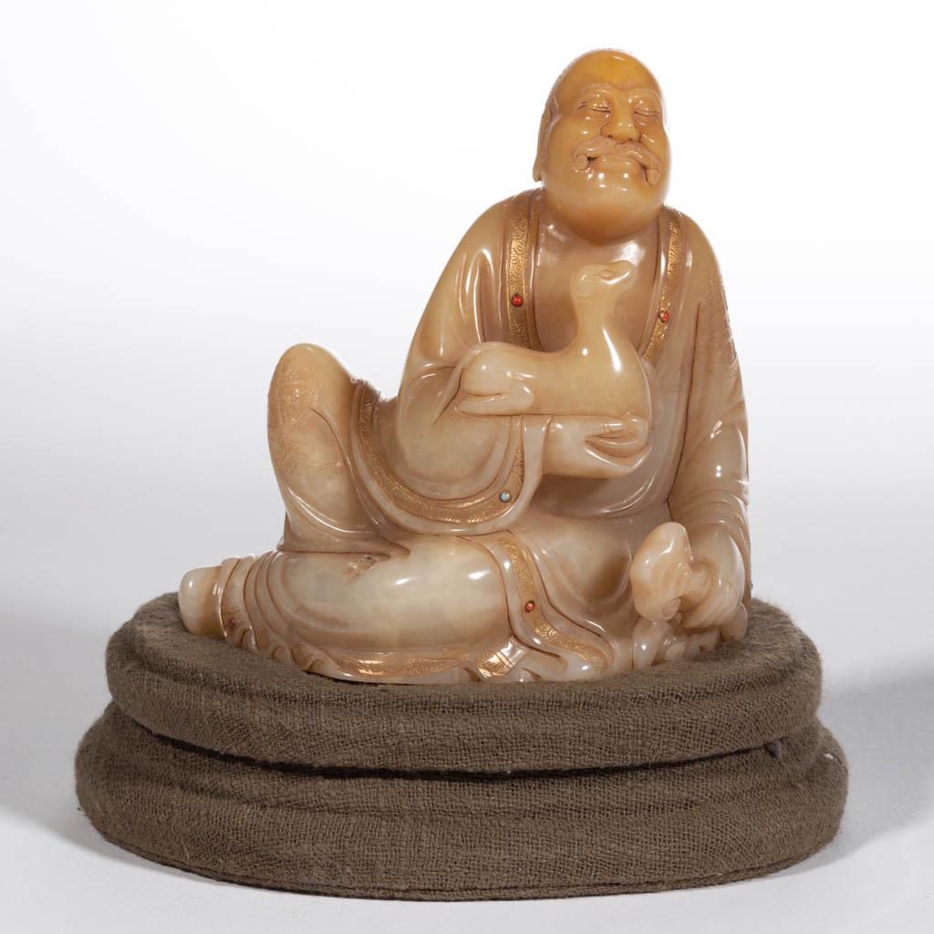 CHINESE SOAPSTONE SHOUSHAN LUOHAN FIGURE: CHINESE SOAPSTONE SHOUSHAN LUOHAN FIGURE, reclining with a smiling, relaxed facial expression, holding a dog in right arm, and a ruyi in left, wearing robe with gilded accents and set with small stone