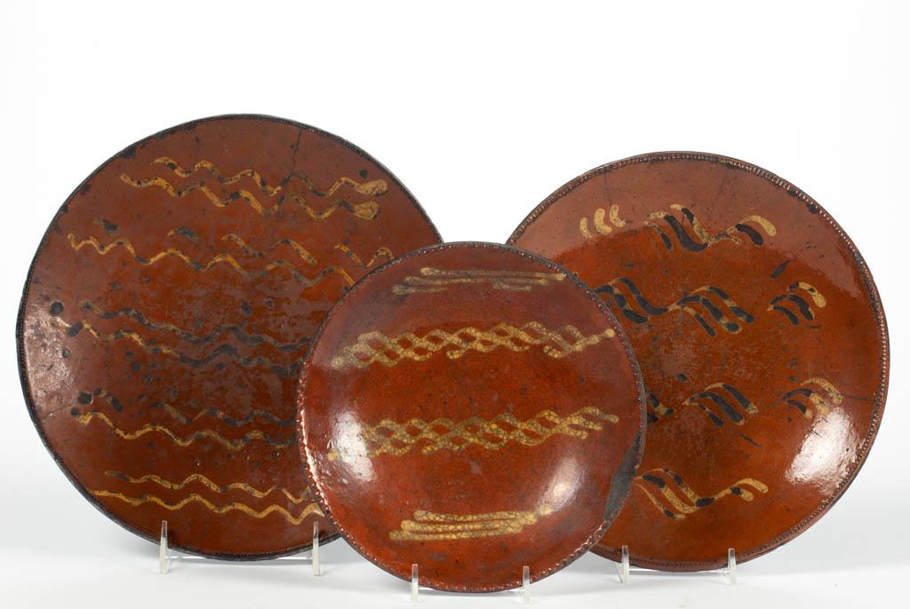AMERICAN DECORATED EARTHENWARE / REDWARE PLATES, LOT OF: AMERICAN DECORATED EARTHENWARE / REDWARE PLATES, LOT OF THREE, lead-glazed interior, each having coggled rim with wavy yellow slip-trailed decorations. First half 19th century. 1 5/8" HOA, 8 1/4" to 1