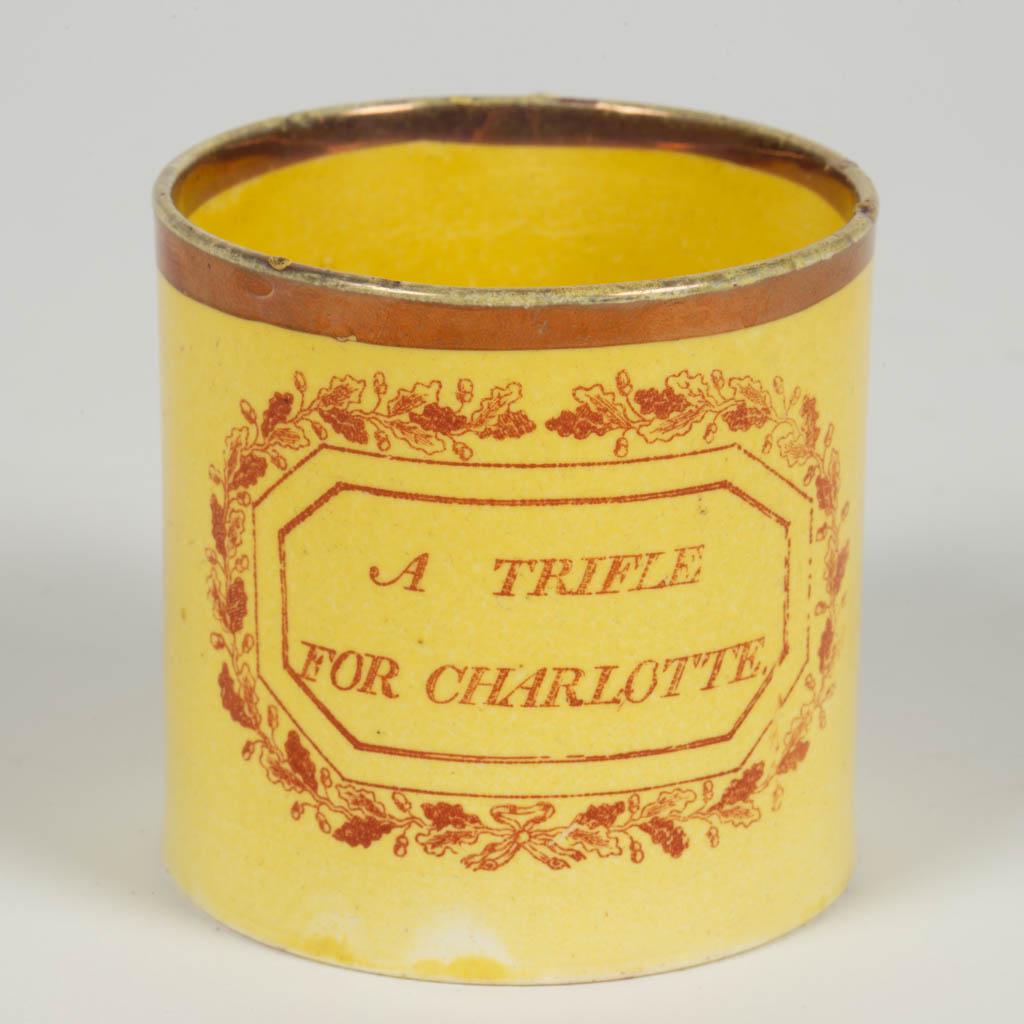 ENGLISH CANARY WARE CERAMIC CHILD'S MUG: ENGLISH CANARY WARE CERAMIC CHILD'S MUG, straight-sided form, featuring red/pink transfer "A TRIFLE / FOR CHARLOTTE" with a band of foliate designs around the wording in canted-corner rectangular brac