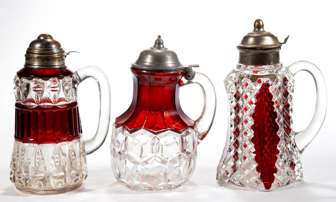 ASSORTED EAPG - RUBY-STAINED SYRUP PITCHERS, LOT OF: ASSORTED EAPG - RUBY-STAINED SYRUP PITCHERS, LOT OF THREE, colorless, comprising a Hexagon Block, a Torpedo, and a Fostoria's Lorraine, each having a period lid. Various makers. Fourth quarter 19th/fi