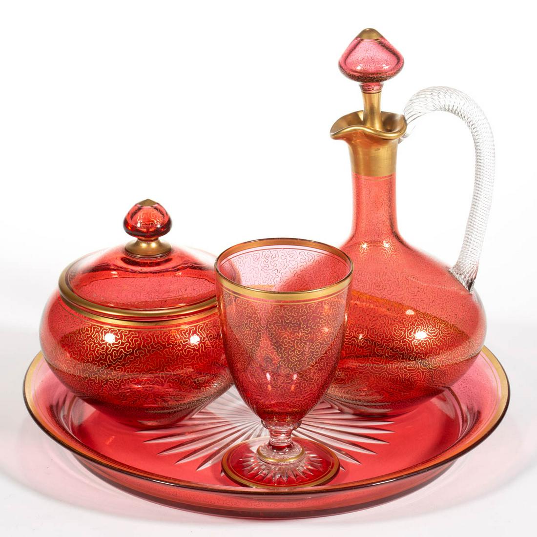 BOHEMIAN GILT-DECORATED ARTICLES, LOT OF FOUR: BOHEMIAN GILT-DECORATED ARTICLES, LOT OF FOUR, cranberry with gilt decoration, comprising a ewer, a covered sugar bowl, a goblet, and a water tray. Probably Moser or Harrach. Late 19th/first quarter 2