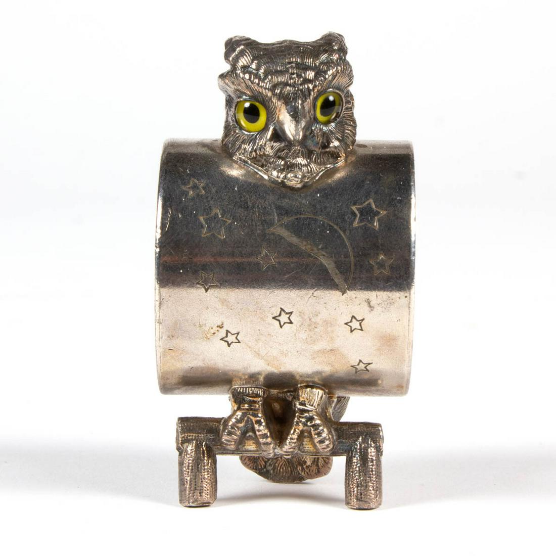 OWL FIGURAL SILVER-PLATED NAPKIN RING: OWL FIGURAL SILVER-PLATED NAPKIN RING, depicting an owl with yellow glass eyes attached to a napkin ring with decoration of the moon and stars to the front and folate motifs surrounding a shield to th