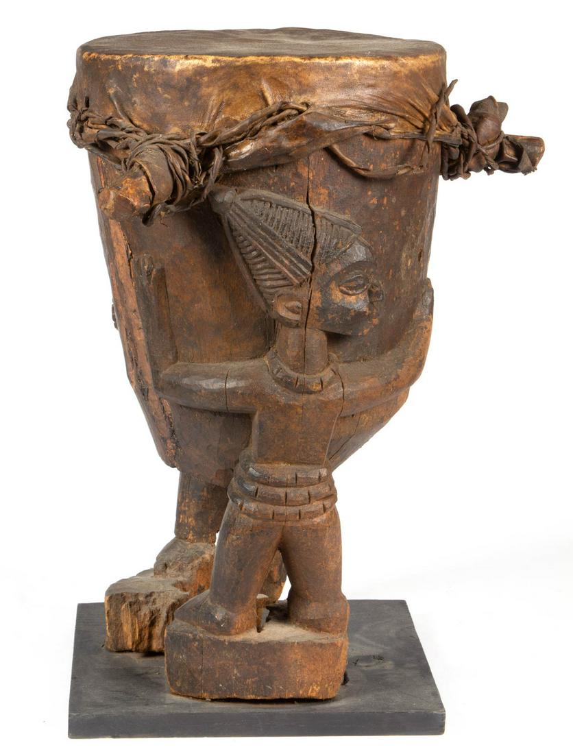 AFRICAN CARVED FIGURAL DRUM: AFRICAN CARVED FIGURAL DRUM, featuring two figures holding up a drum with likely original cover, applied to a later base. Old surface with warm color. 20th century. 20" HOA, 11" D top. As-found condit