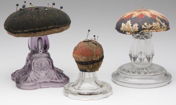 THREE MAKE-DO PRESSED GLASS BASE PINCUSHIONS: THREE MAKE-DO PRESSED GLASS BASE PINCUSHIONS, comprised of two lamp bases, and one compote or cake stand base, each fitted with a pinchusion top. 19th century. 4" to 6" h. Provenance: From a North Car