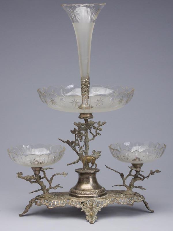 SILVER-PLATED AND CUT GLASS EPERGNE: SILVER-PLATED AND CUT GLASS EPERGNE, three cut and frosted colorless glass bowls and single vase, raised on an ornate bolted stand decorated with a stag, three trees, and cupid masks. First quarter 20