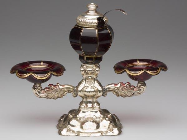 CASED RUBY GLASS CONDIMENT SET: CASED RUBY GLASS CONDIMENT SET, comprised of two salt cellars and a central mustard pot mounted on a silver-like weighted stand, pot with original cover and ladle, glass elements with original gilding