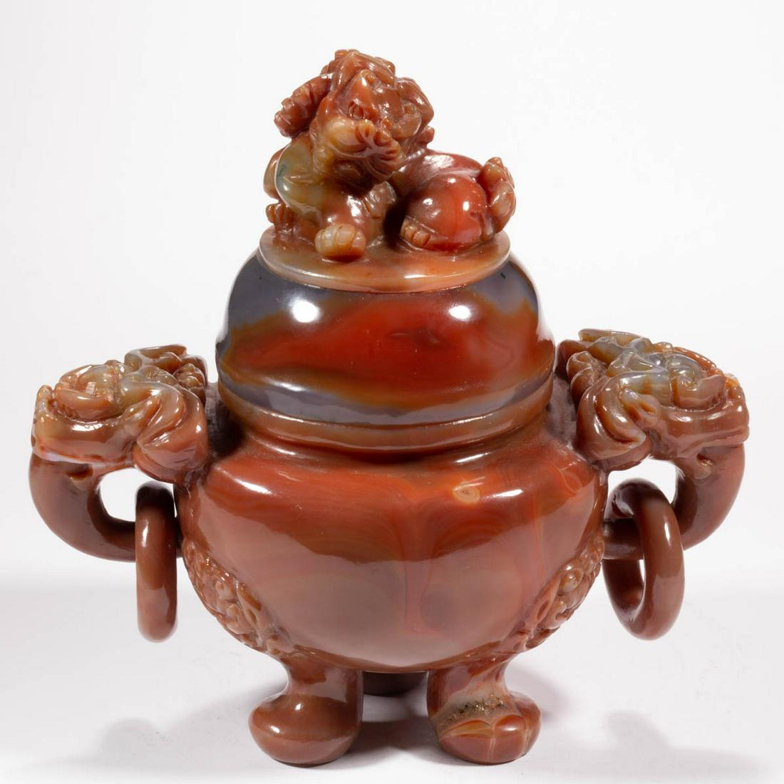CHINESE CARVED RED AGATE CENSER: CHINESE CARVED RED AGATE CENSER, having a round well, featuring carved guardian lion handles with attached rings, raised on three pad feet with lion/dragon head knees, topped by a dome cover with figu