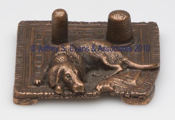 BRONZE FIGURAL THIMBLE HOLDER AND SPOOL STAND: BRONZE FIGURAL THIMBLE HOLDER AND SPOOL STAND, depicting a large dog lying on a rug, underside impressed "R" within a circle and "56". Late 19th/early 20th century. 4 1/8" h overall, 2 1/4" x 3 1/4".