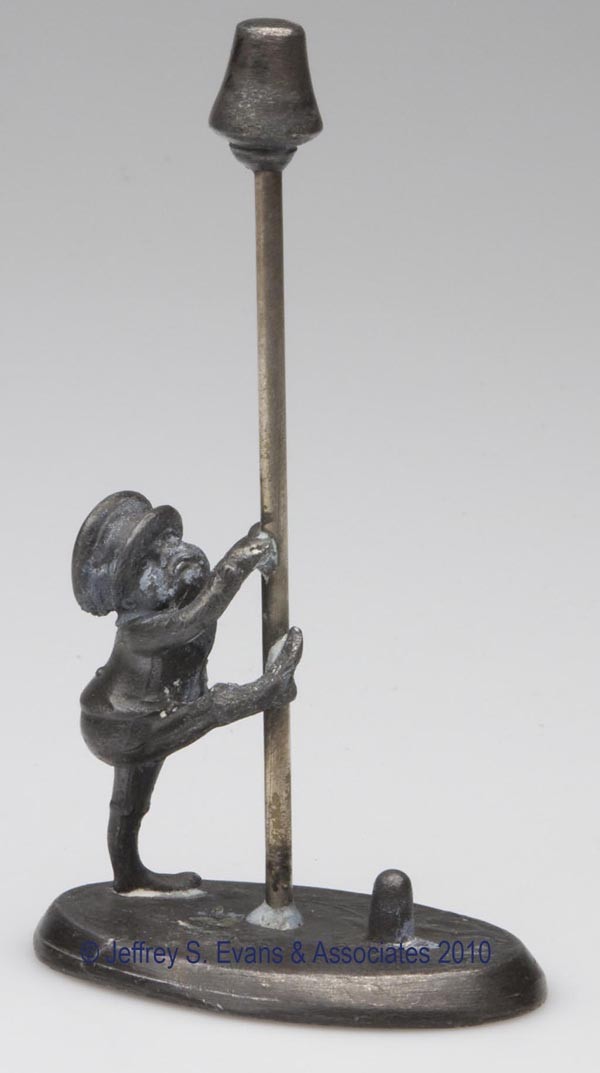 FIGURAL WHITE METAL THIMBLE HOLDER AND SPOOL STAND: FIGURAL WHITE METAL THIMBLE HOLDER AND SPOOL STAND, depicting a brownie climbing a pole, oval base with embossed leaf decoration to top, no markings. Late 19th/early 20th century. 4 1/2" h overall, 1 