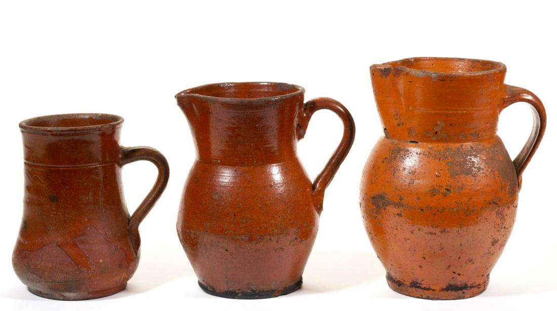 AMERICAN EARTHENWARE / REDWARE VESSELS, LOT OF THREE: AMERICAN EARTHENWARE / REDWARE VESSELS, LOT OF THREE, lead-glazed, comprising a low-belly mug with incised sine wave below the neck ring, and two ovoid-form pitchers, each with ridged, strap handle. 1