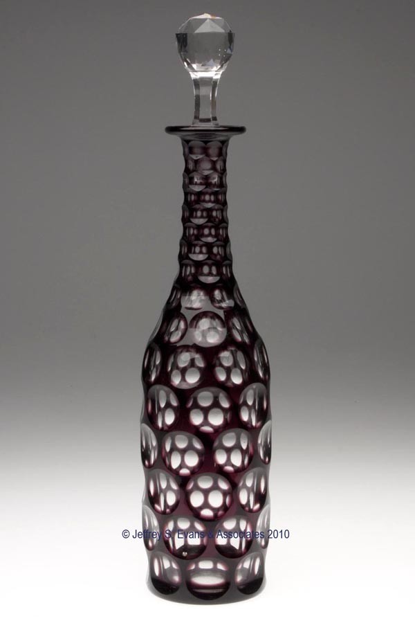 CUT OVERLAY PUNTY DECANTER: CUT OVERLAY PUNTY DECANTER, deep amethyst to colorless, cylindrical form with tall neck featuring all-over cutting, plain base with wide polished pontil mark, well-fitting colorless facet-cut stopper.