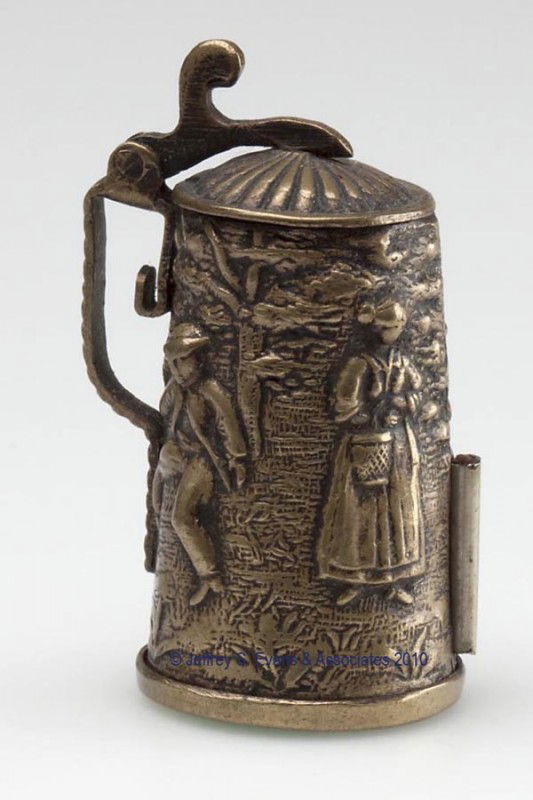 BRASS FIGURAL TAPE MEASURE: BRASS FIGURAL TAPE MEASURE, depicting a stein with molded figures of men and woman, top hinges open to a compartment for a thimble, disk bottom revolves for winding tape, silk tape calibrated in centi