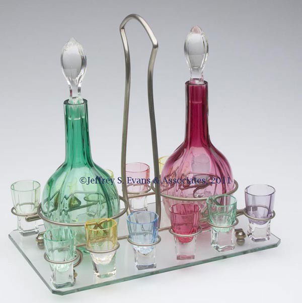 EUROPEAN COLORED GLASS LIQUOR SET: EUROPEAN COLORED GLASS LIQUOR SET, comprising two decanters and 12 shot glasses, each molded with four ribs and featuring a polished base, all fitted in a glass and nickel-plated stand. Probably Czech