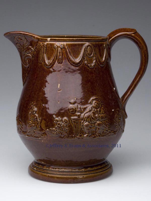 SIGNED "JORDAN" ROCKINGHAM-STYLE PITCHER: SIGNED "JORDAN" ROCKINGHAM-STYLE PITCHER, brown glaze, embossed scene with heavy-set drinking man in a classical garden, rim surrounded by a swag with bowknots and medallions, base impressed "JORDAN".