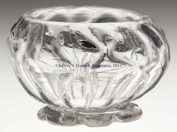 PATTERN-MOLDED FOOTED SALT: PATTERN-MOLDED FOOTED SALT, colorless, compressed globular-form bowl molded with 16 nipped ribs, raised on a tooled seven-petal low foot with a rough pontil mark. Probably England. 1780-1820. 1 5/8" H