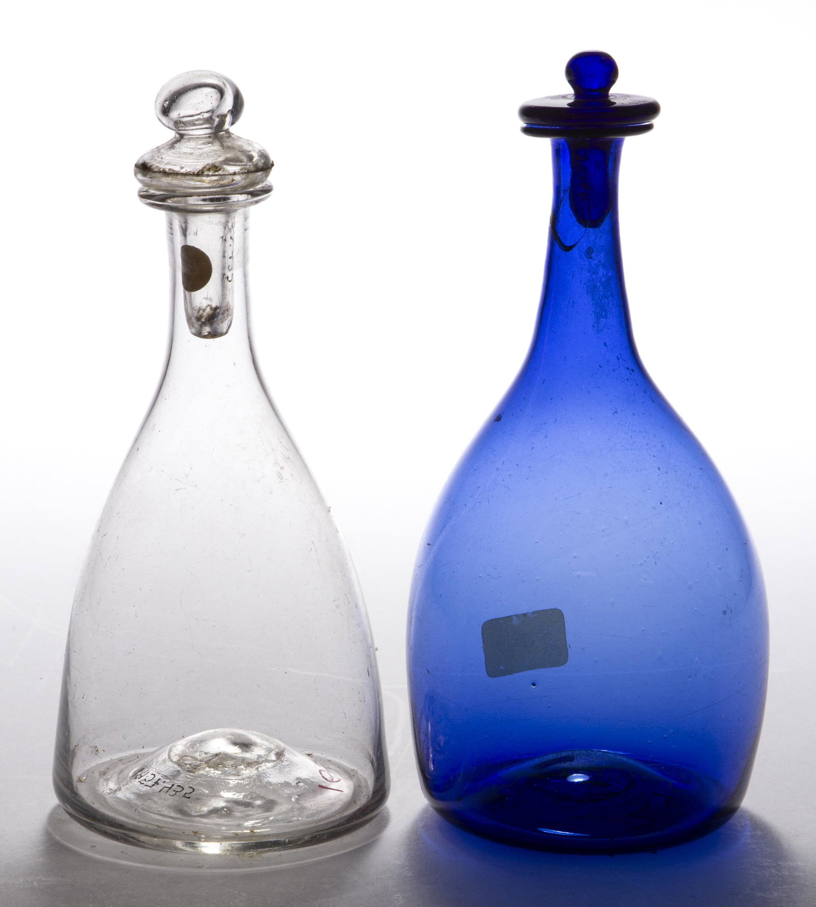 FREE-BLOWN TOILET BOTTLES, LOT OF TWO: FREE-BLOWN TOILET BOTTLES, LOT OF TWO, colorless and medium blue, each globular/tapered form with a matching tam-o-shanter stopper, rough pontil marks. Various makers. First half 19th century. 6 1/4" 