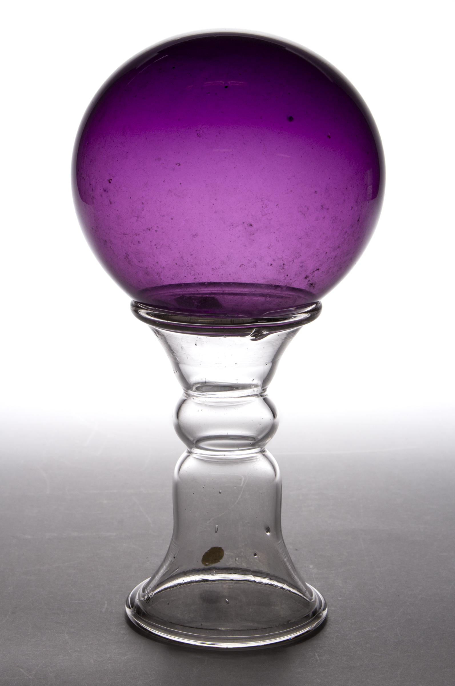 FREE-BLOWN GLASS WITCH BALL AND STAND: FREE-BLOWN GLASS WITCH BALL AND STAND, comprising an amethyst ball resting on a colorless waisted stand. Maker unverified. 19th century. Ball: 4 1/4" D. Stand: 5" H, 2 7/8" D rim, 3 1/8" D base. Publi