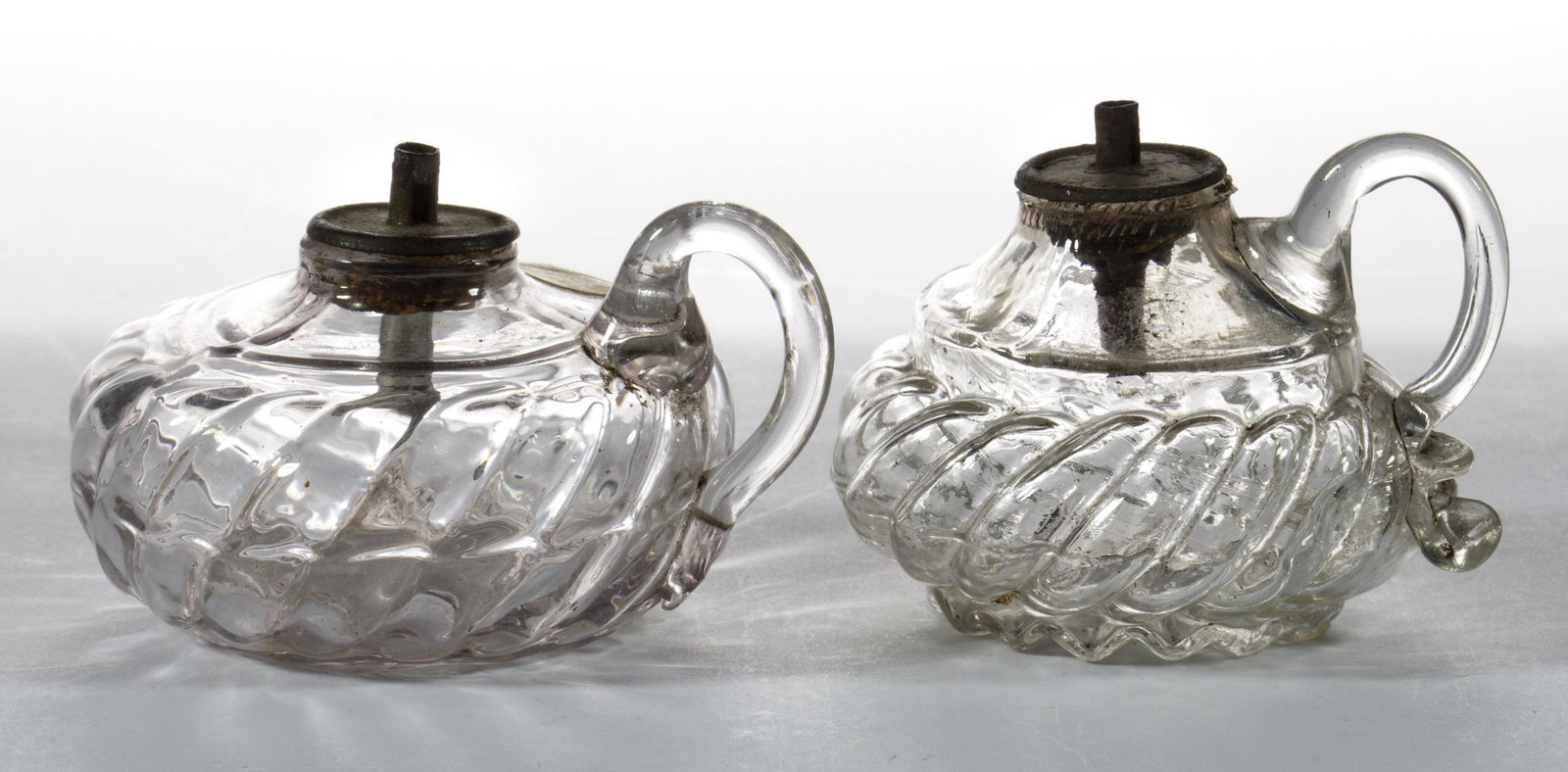 BLOWN-MOLDED WHALE OIL TOY FINGER / SPARKING LAMPS, LOT OF TWO: BLOWN-MOLDED WHALE OIL TOY FINGER / SPARKING LAMPS, LOT OF TWO, colorless, each with swirled ribs, delicate applied handles, and rayed bases with rough pontil mark. Each fitted with a tin single-tube 