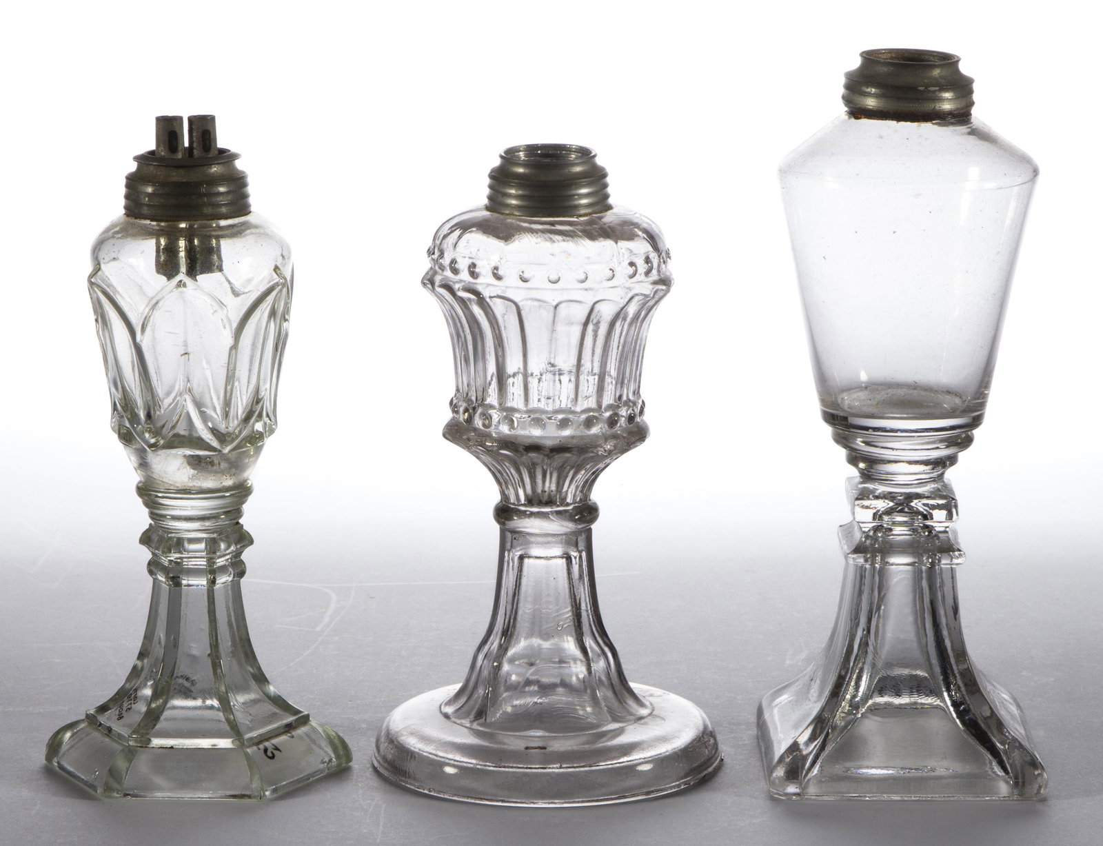 ASSORTED PRESSED AND BLOWN GLASS WHALE OIL / FLUID STAND LAMPS, LOT OF THREE: ASSORTED PRESSED AND BLOWN GLASS WHALE OIL / FLUID STAND LAMPS, LOT OF THREE, colorless, comprising an unpatterned example on a flaring square base, a Tulip, and a Beaded Flutes example, each having a