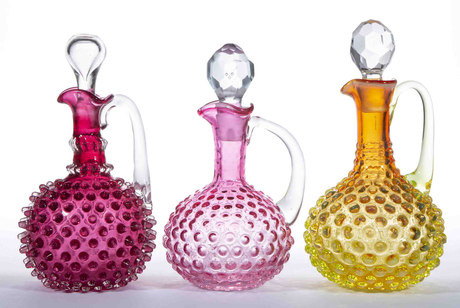 ASSORTED HOBNAIL / DEW DROP GLASS CRUETS, LOT OF THREE,: ASSORTED HOBNAIL / DEW DROP GLASS CRUETS, LOT OF THREE, comprising two cased cranberry examples, and one having an amberina-type, each having a period stopper and applied handle. Various makers. Late 