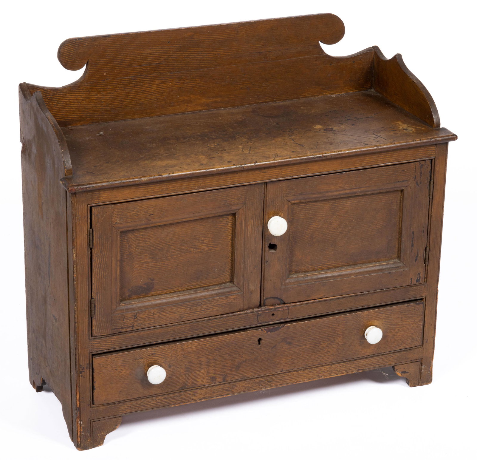 AMERICAN PAINT-DECORATED PINE CHILD'S CUPBOARD,: featuring applied gallery above two hinged fielded panel doors concealing an open interior over full-length drawer with likely original porcelain knobs, raised on cut-out feet. Retains original grain-