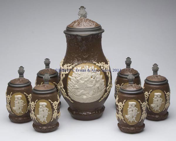 METTLACH #1028 CERAMIC SEVEN-PIECE STEIN SET: METTLACH #1028 CERAMIC SEVEN-PIECE STEIN SET, comprising a 4.0L master jug and six .5L steins, each relief molded and inscribed, and with an inlay lid and full base markings. Early 20th century. 13 1/