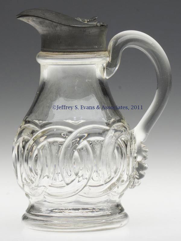 BLOWN-MOLDED WEDDING RING MOLASSES CAN / JUG: BLOWN-MOLDED WEDDING RING MOLASSES CAN / JUG, colorless, small pyriform body with an applied solid handle, raised on a pronounced foot with a plain base. Original Britannia hinged lid. Probably Pittsb