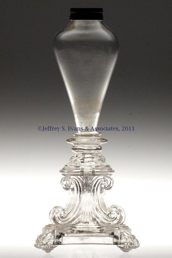 FREE-BLOWN AND PRESSED WHALE OIL STAND LAMP: FREE-BLOWN AND PRESSED WHALE OIL STAND LAMP, colorless, bulb font raised on a complex triangular C-scroll base terminating in paw feet, bladed wafer construction. Factory ground under two feet to leve