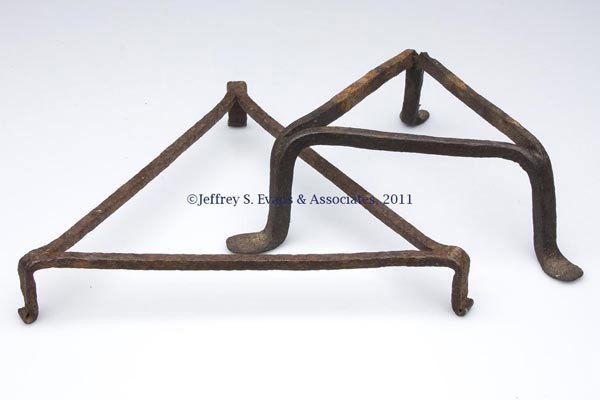 TWO WROUGHT-IRON TRIANGULAR TRIVETS: TWO WROUGHT-IRON TRIANGULAR TRIVETS, one with pad feet, the other curled. 18th or 19th century. 3" H, 8" LOA, and 1 1/4" H, 10 3/4" LOA. Provenance: Estate collection of Dorothy and Marshall Jenkins.