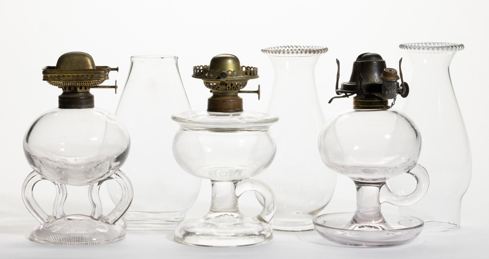 ASSORTED PATTERN KEROSENE FOOTED FINGER LAMPS, LOT OF THREE: ASSORTED PATTERN KEROSENE FOOTED FINGER LAMPS, LOT OF THREE,colorless,each with a molded handle, patterns include a Ripley with double handles and a solid glass stem; a Lomax Oil Guard example; and a 