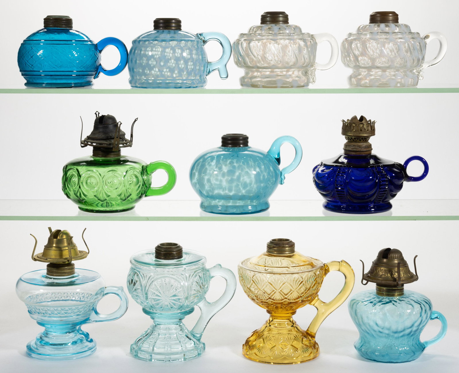 ASSORTED GLASS KEROSENE FINGER LAMPS, LOT OF 11: ASSORTED GLASS KEROSENE FINGER LAMPS, LOT OF 11, various colors,including two Markham Swirls, a Coolidge Drape, a Windows, and blue with opal spatter, each having a brass collar. Four fitted with a pe