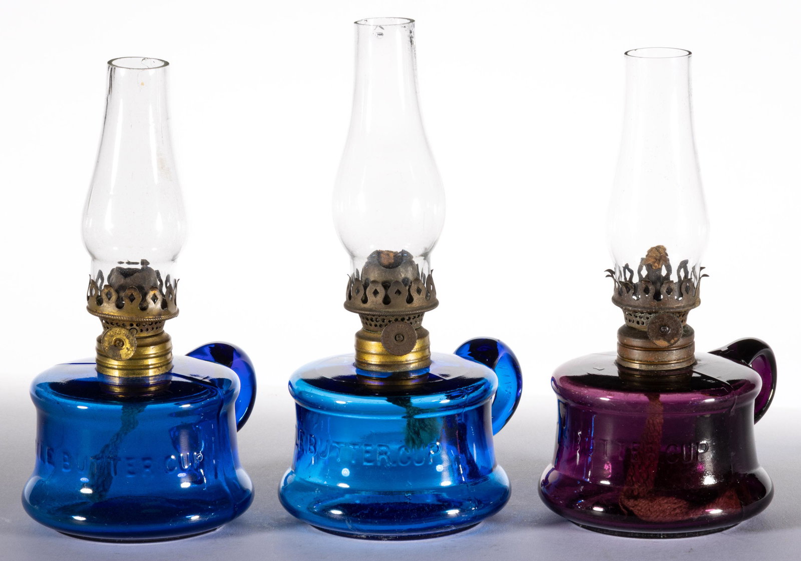 ASSORTED LITTLE BUTTERCUP MINIATURE FINGER LAMPS, LOT OF THREE: ASSORTED LITTLE BUTTERCUP MINIATURE FINGER LAMPS, LOT OF THREE,deep amethyst, and sapphire blue,each with an applied handle, concave fonts embossed "LITTLE BUTTERCUP", period collars. Each fitted with