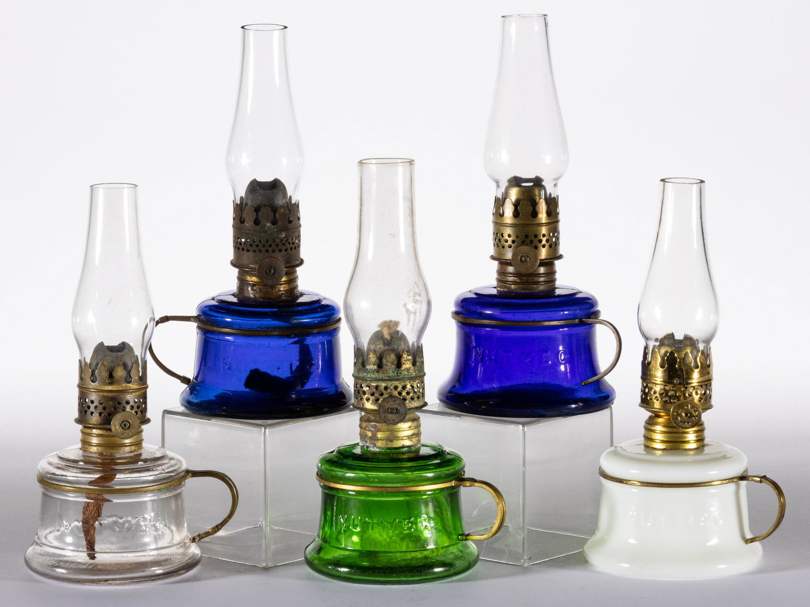 ASSORTED NUTMEG MINIATURE FINGER LAMPS, LOT OF FIVE: ASSORTED NUTMEG MINIATURE FINGER LAMPS, LOT OF FIVE,cobalt blue, colorless, green, and opaque white,each embossed "NUTMEG" on font, original brass wire handle. Each fitted with a burner, colorless chi