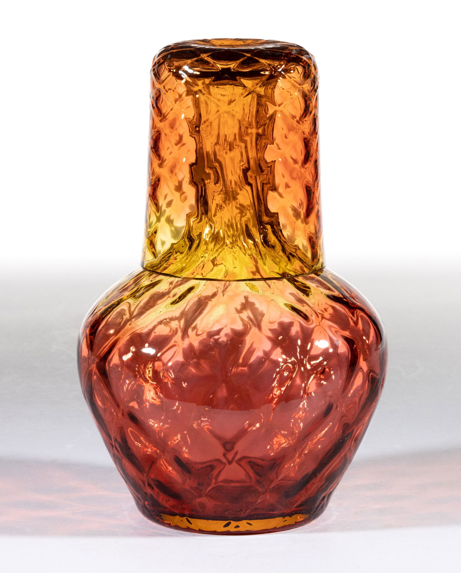 VICTORIAN AMBERINA DIAMOND-QUILT GLASS TUMBLE-UP: VICTORIAN AMBERINA DIAMOND-QUILT GLASS TUMBLE-UP, amberina, globular form bottle with a matching tumbler. Maker unverified. Fourth quarter 19th century. 7 1/4" HOA, 6 7/8" H bottle. </