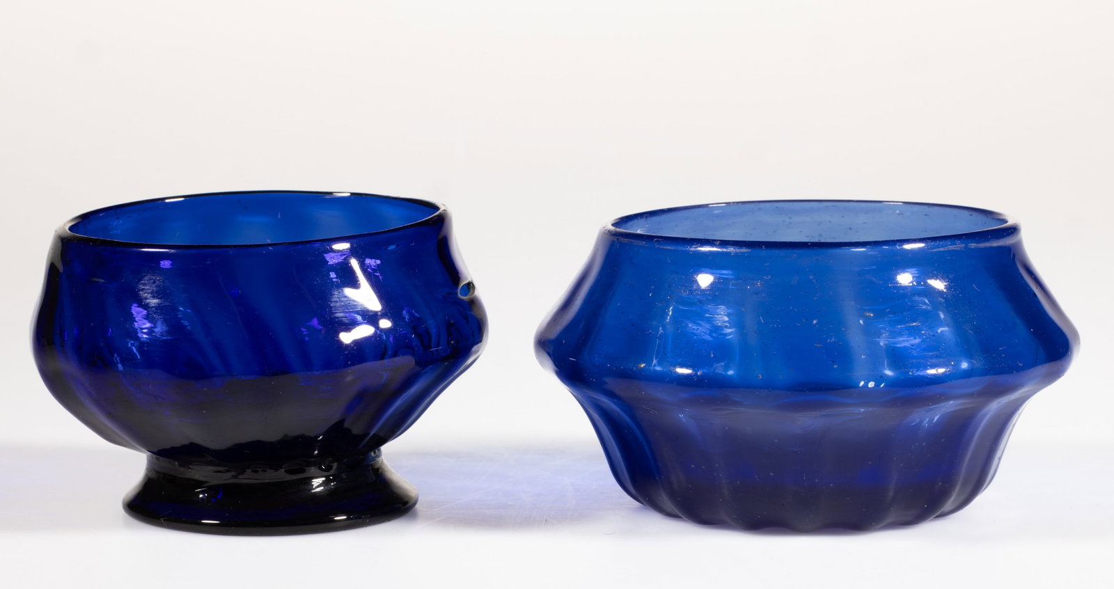 PATTERN-MOLDED GLASS OPEN SALTS, LOT OF TWO: PATTERN-MOLDED GLASS OPEN SALTS, LOT OF TWO, medium and deep blue, comprising one compressed form with 17 ribs, and one having an applied foot with 16 ribs, rough pontil marks. Probably English. 1810-